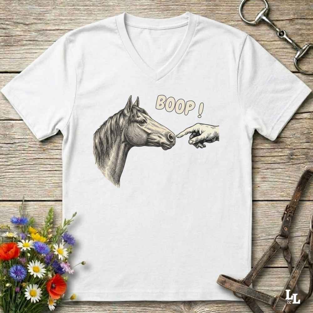 A white V-neck t-shirt laid flat on a rustic wooden surface, featuring a graphic of a horse's head and a hand pointing at it with the text 'BOOP!'. The shirt is styled with equestrian hardware and flowers.