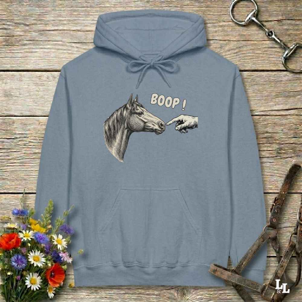 A dusty blue hoodie laid flat on a rustic wooden surface, featuring a graphic of a horse head and a pointing hand with the text 'BOOP!'. The hoodie is styled with equestrian-themed props including a halter, tack, and a small bouquet of wildflowers.