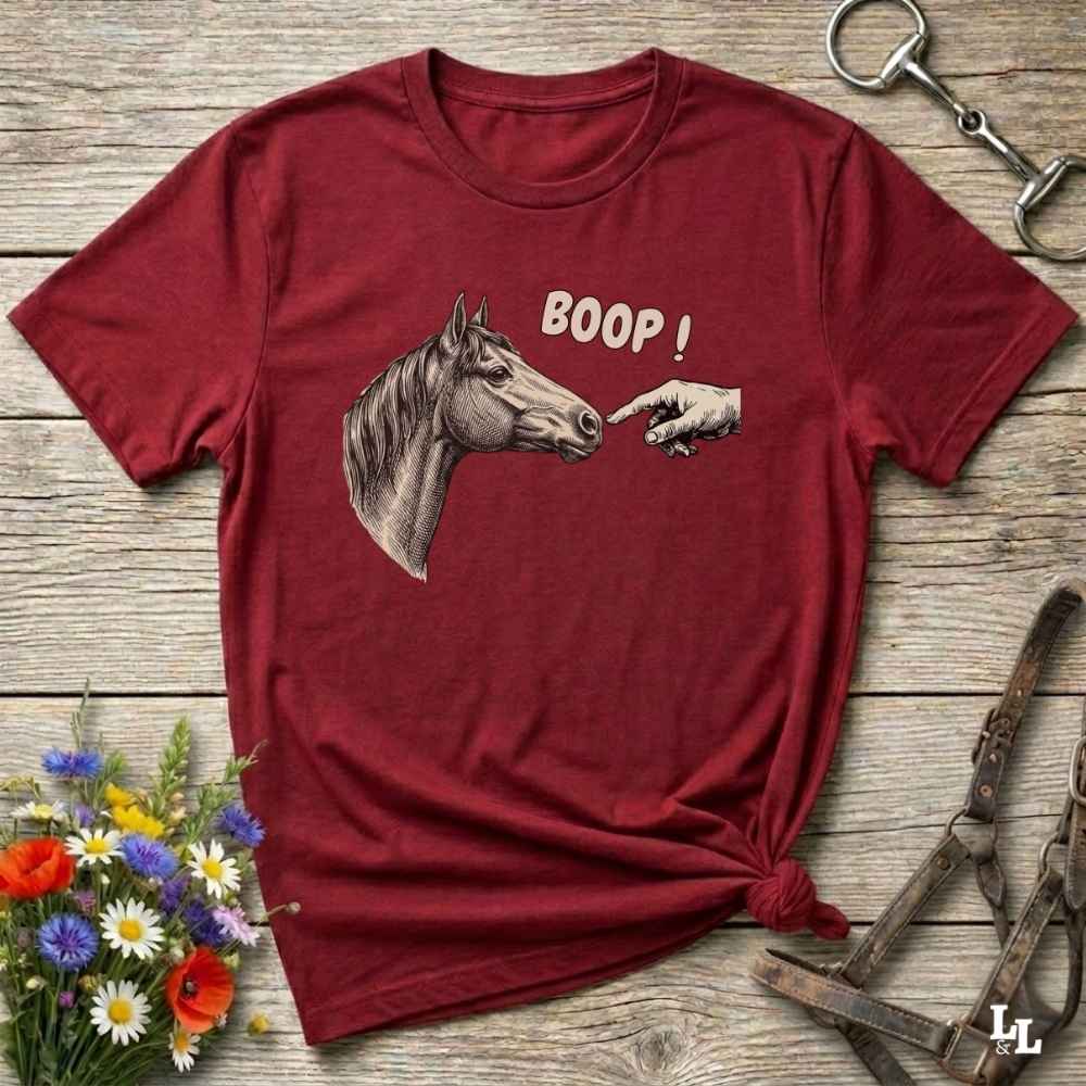 Red t-shirt with horse and hand graphic and 'BOOP!' text on a wooden surface with flowers and equestrian equipment.