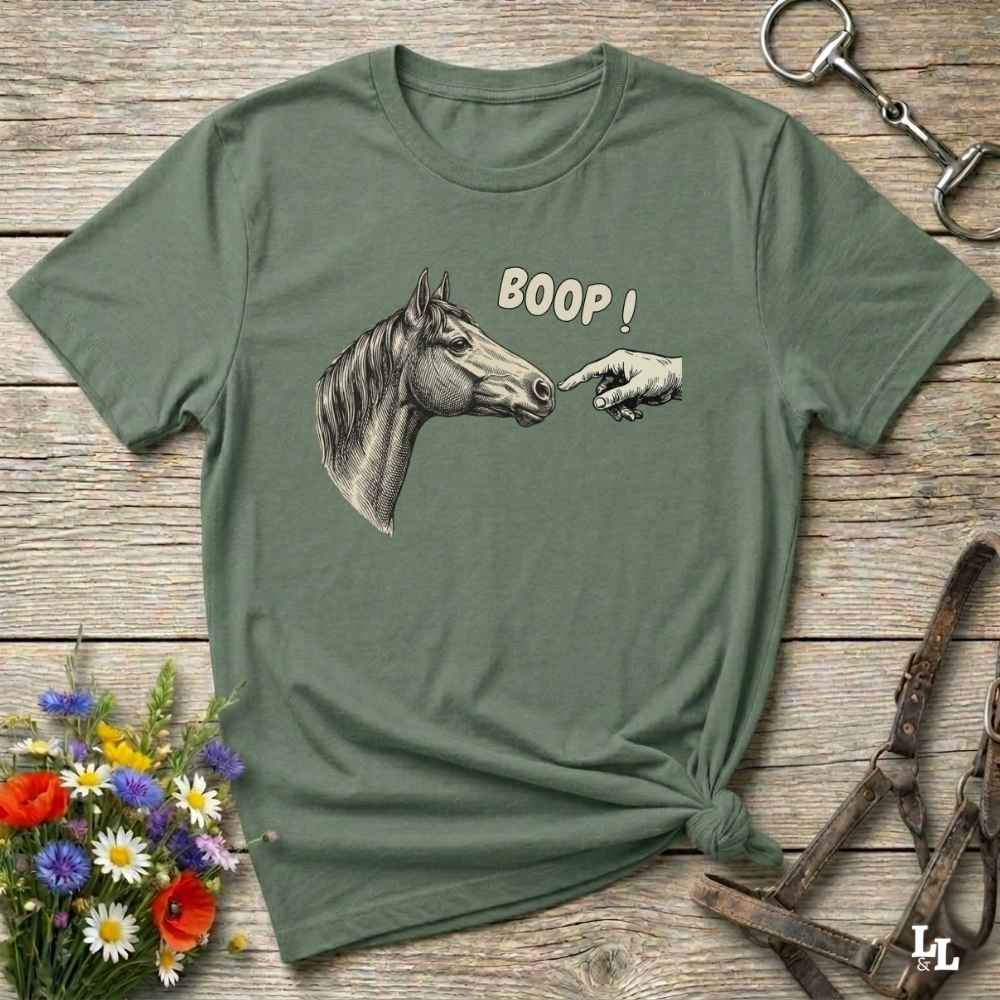 Green t-shirt with horse and hand graphic and 'BOOP!' text on a wooden surface with flowers and equestrian equipment.