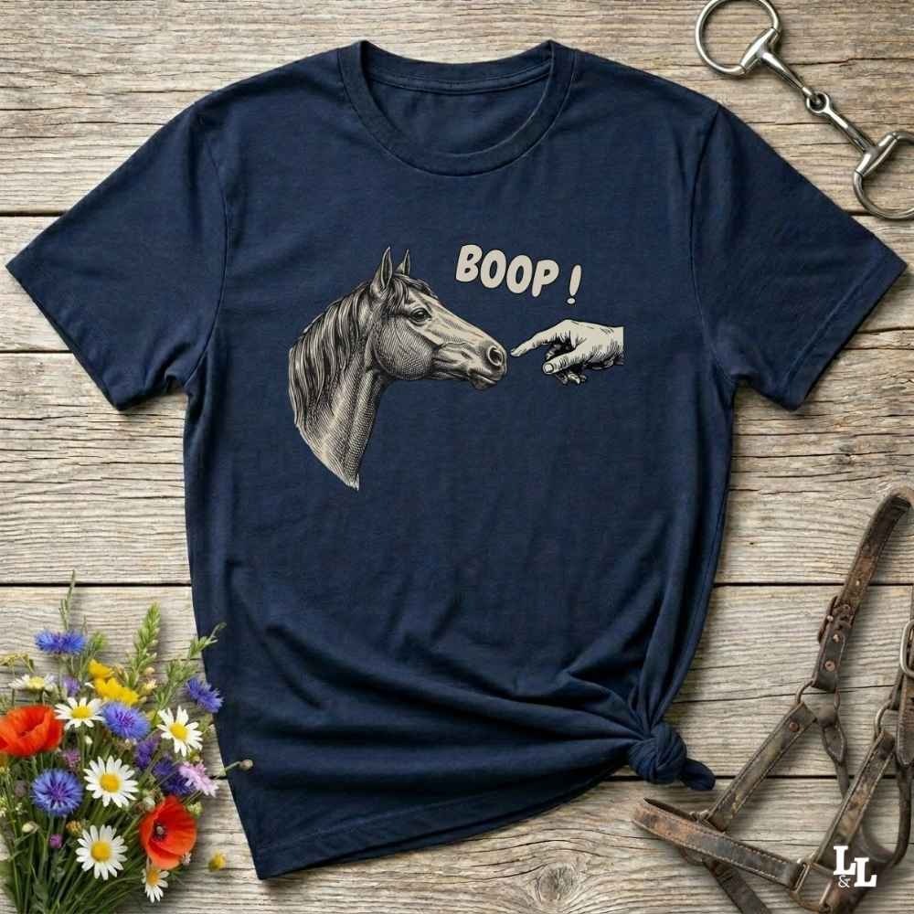 Navy blue t-shirt with horse graphic and 'BOOP!' text on a wooden surface with flowers and equestrian tools.