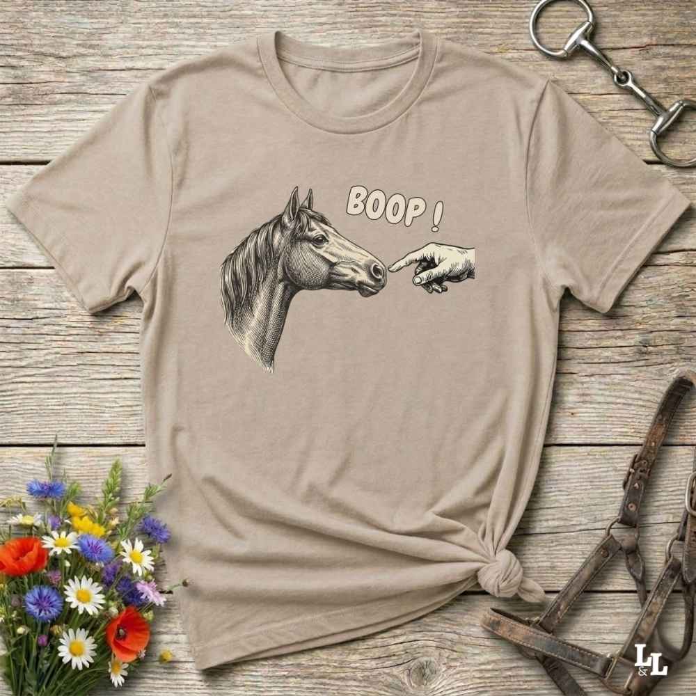 Beige t-shirt with horse graphic and 'BOOP!' text on a wooden surface with flowers and equestrian tools.