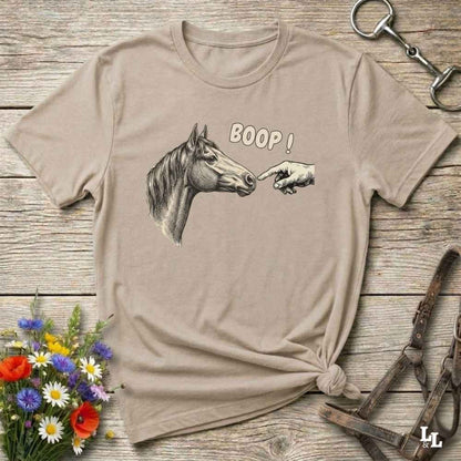 Beige t-shirt with horse graphic and 'BOOP!' text on a wooden surface with flowers and equestrian tools.
