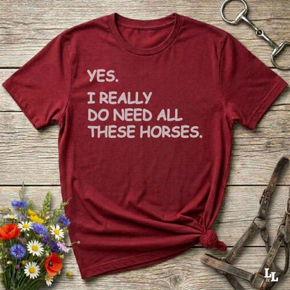 Red t-shirt with text 'YES. I REALLY DO NEED ALL THESE HORSES.' on a wooden surface with flowers and horse equipment.