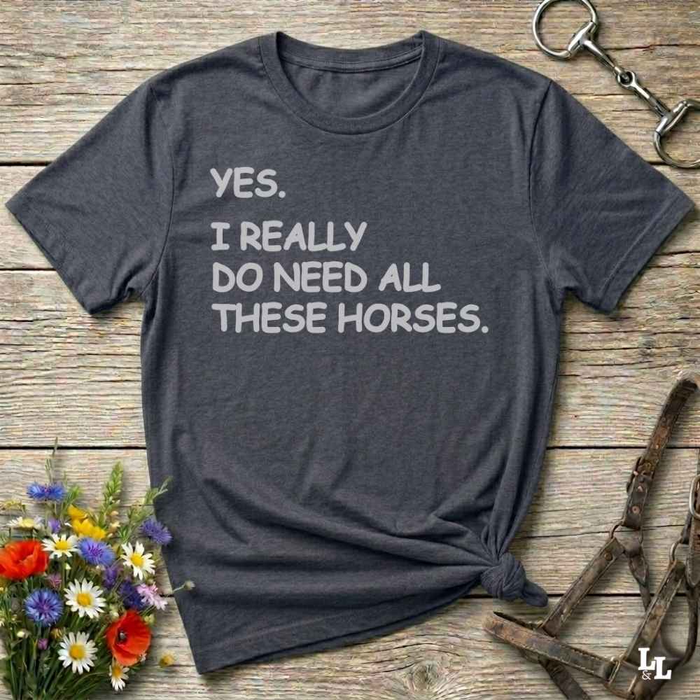 Gray t-shirt with text 'YES. I REALLY DO NEED ALL THESE HORSES.' on a wooden surface with floral and equestrian accessories.