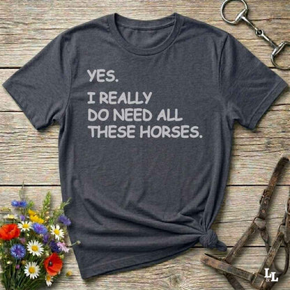 Gray t-shirt with text 'YES. I REALLY DO NEED ALL THESE HORSES.' on a wooden surface with floral and equestrian accessories.