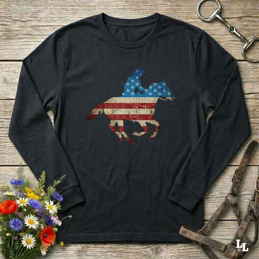 Black long-sleeve shirt with American flag horse design on a wooden surface with flowers and equestrian equipment.
