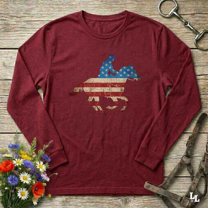 Red long-sleeve shirt with American flag and horse graphic on a wooden surface with flowers and equestrian equipment.