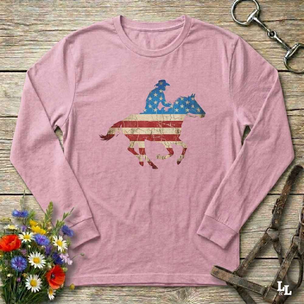 Pink long-sleeve shirt with a horse and rider design featuring an American flag on a wooden surface with floral and equestrian elements.