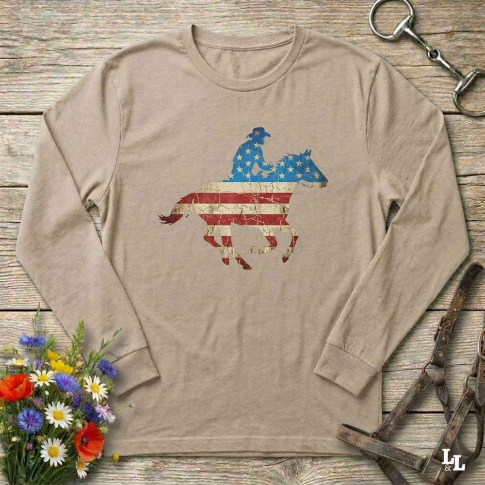 Beige long-sleeve shirt with a graphic of a horse and rider on an American flag design, placed on a wooden surface with floral and equestrian-themed accessories.