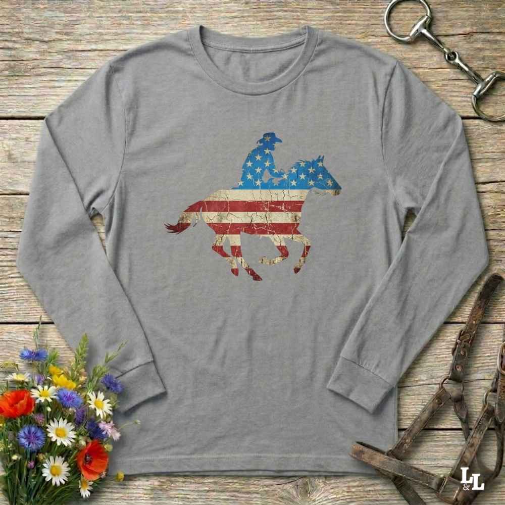 Gray long-sleeve shirt with American flag horse and rider design on a wooden surface with flowers and horse equipment.