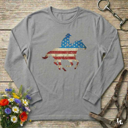 Gray long-sleeve shirt with American flag horse and rider design on a wooden surface with flowers and horse equipment.