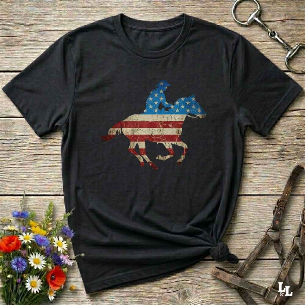 Black t-shirt with American flag and horse design on a wooden surface with flowers and equestrian equipment.