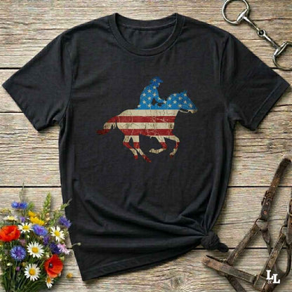 Black t-shirt with American flag and horse design on a wooden surface with flowers and equestrian equipment.