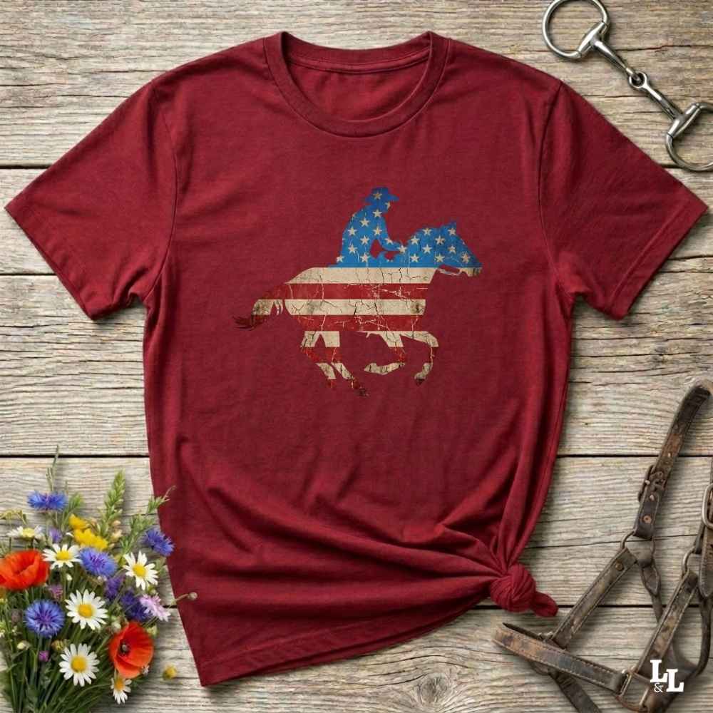 Red t-shirt with a graphic design on a wooden surface with flowers and horse equipment.