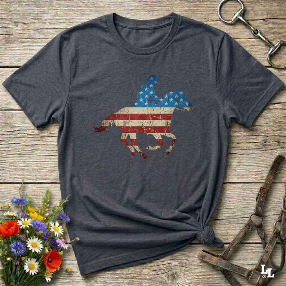 Gray t-shirt with American flag horse design on a wooden surface with flowers and equestrian equipment.