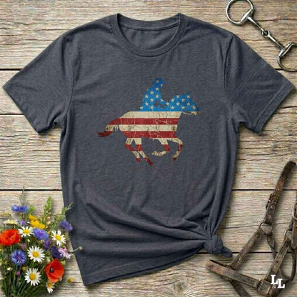 Gray t-shirt with American flag horse design on a wooden surface with flowers and equestrian equipment.