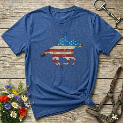 Blue t-shirt with American flag and horse design on a wooden surface with flowers and equestrian equipment.