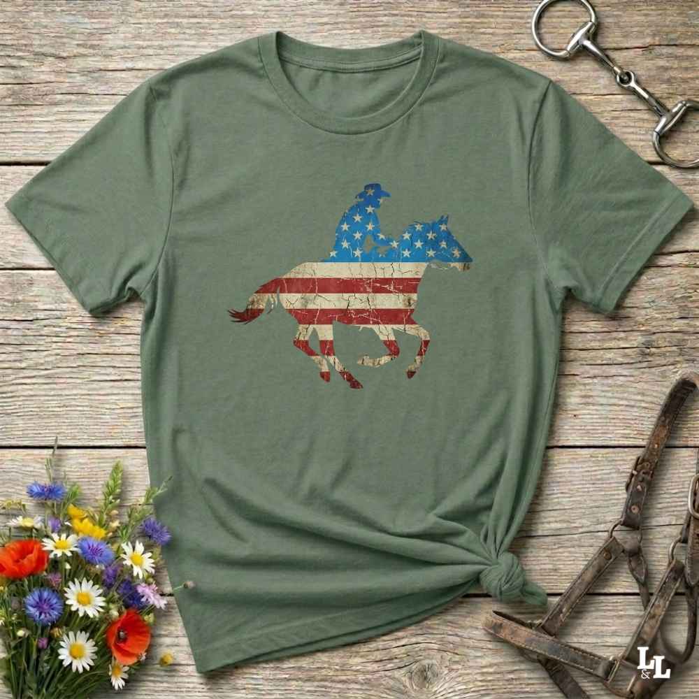 Green t-shirt with American flag horse design on a wooden surface with flowers and equestrian equipment.