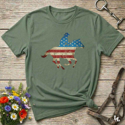Green t-shirt with American flag horse design on a wooden surface with flowers and equestrian equipment.
