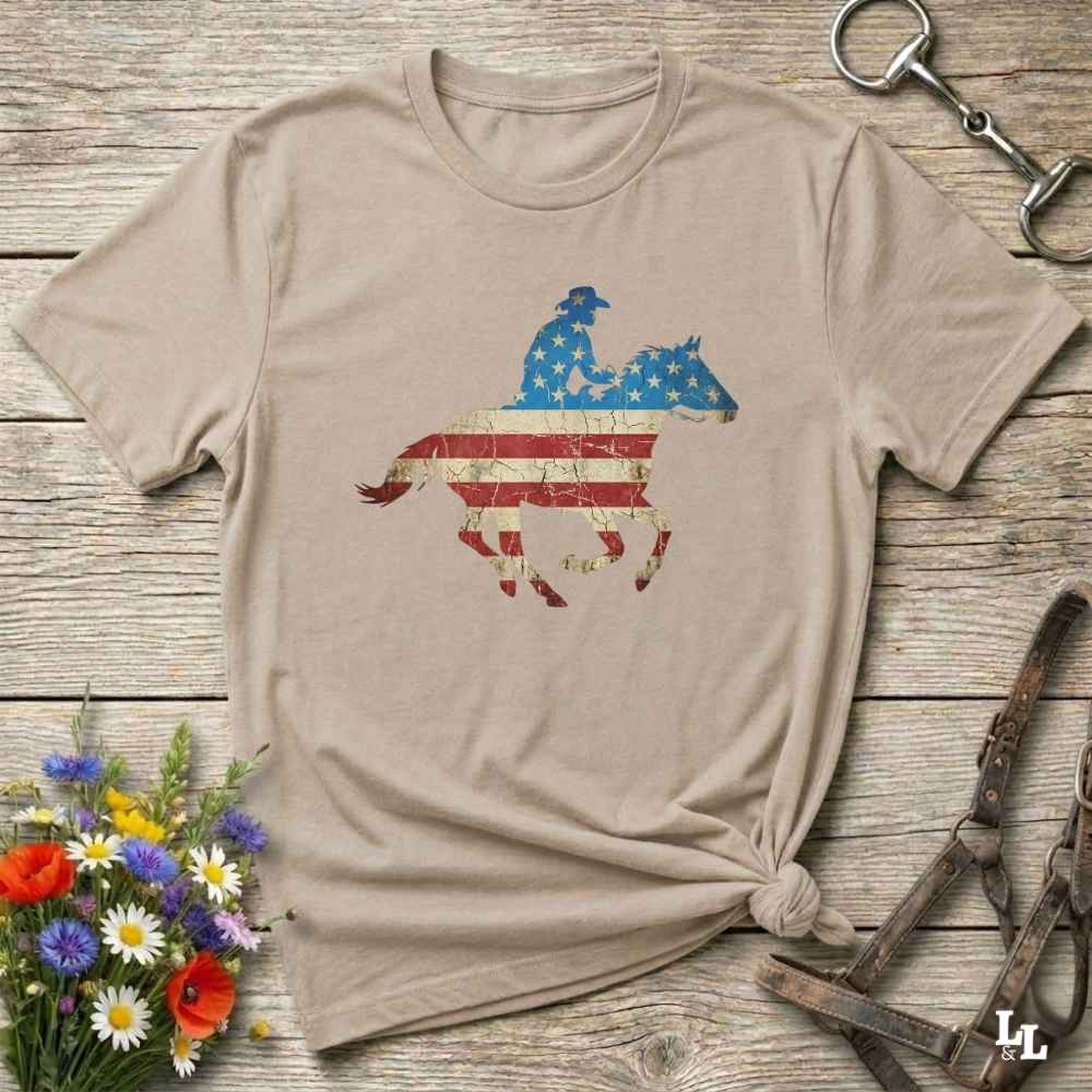 Beige t-shirt with American flag horse and rider graphic on a wooden surface with flowers and equestrian tools.