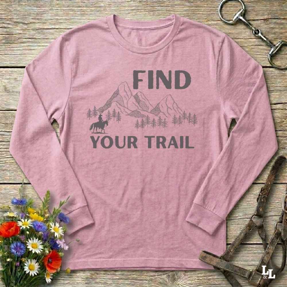 Pink long-sleeve shirt with 'Find Your Trail' graphic on a wooden surface with flowers and horse equipment.