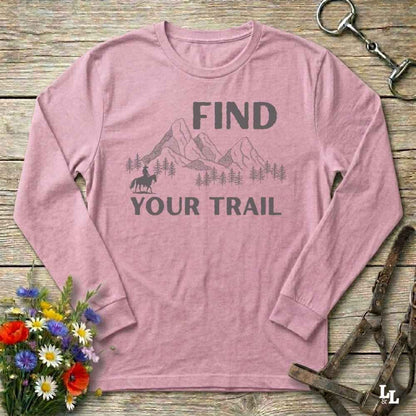Pink long-sleeve shirt with 'Find Your Trail' graphic on a wooden surface with flowers and horse equipment.
