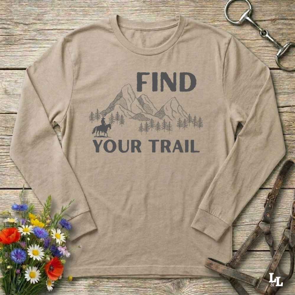 Beige long-sleeve shirt with mountain and horse graphic and 'Find Your Trail' text on a wooden background with flowers and horse equipment.