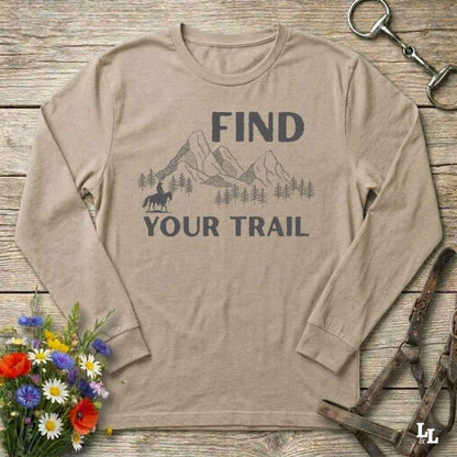 Beige long-sleeve shirt with mountain and horse graphic and 'Find Your Trail' text on a wooden background with flowers and horse equipment.