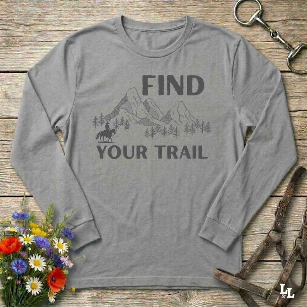 Gray long-sleeve shirt with 'Find Your Trail' graphic on a wooden background with flowers and horse equipment.