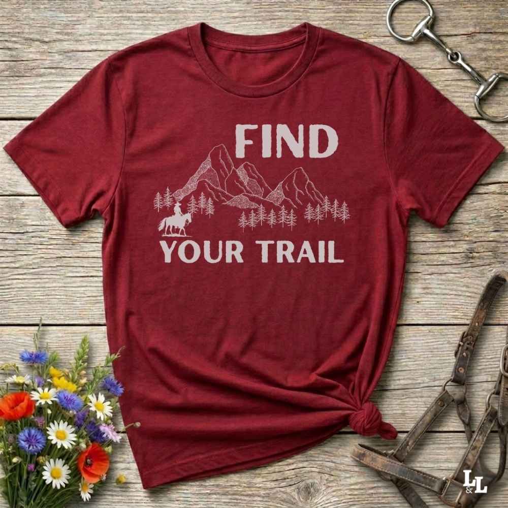 Red t-shirt with 'Find Your Trail' graphic on a wooden surface with flowers and horse equipment.
