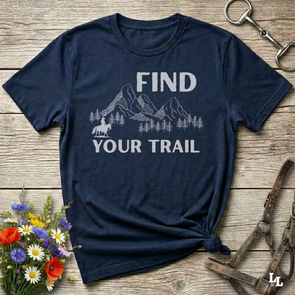 Navy blue t-shirt with 'Find Your Trail' graphic on a wooden surface with flowers and horse equipment.