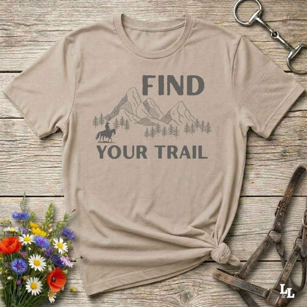 Beige t-shirt with 'Find Your Trail' graphic on a wooden surface with flowers and horse equipment.