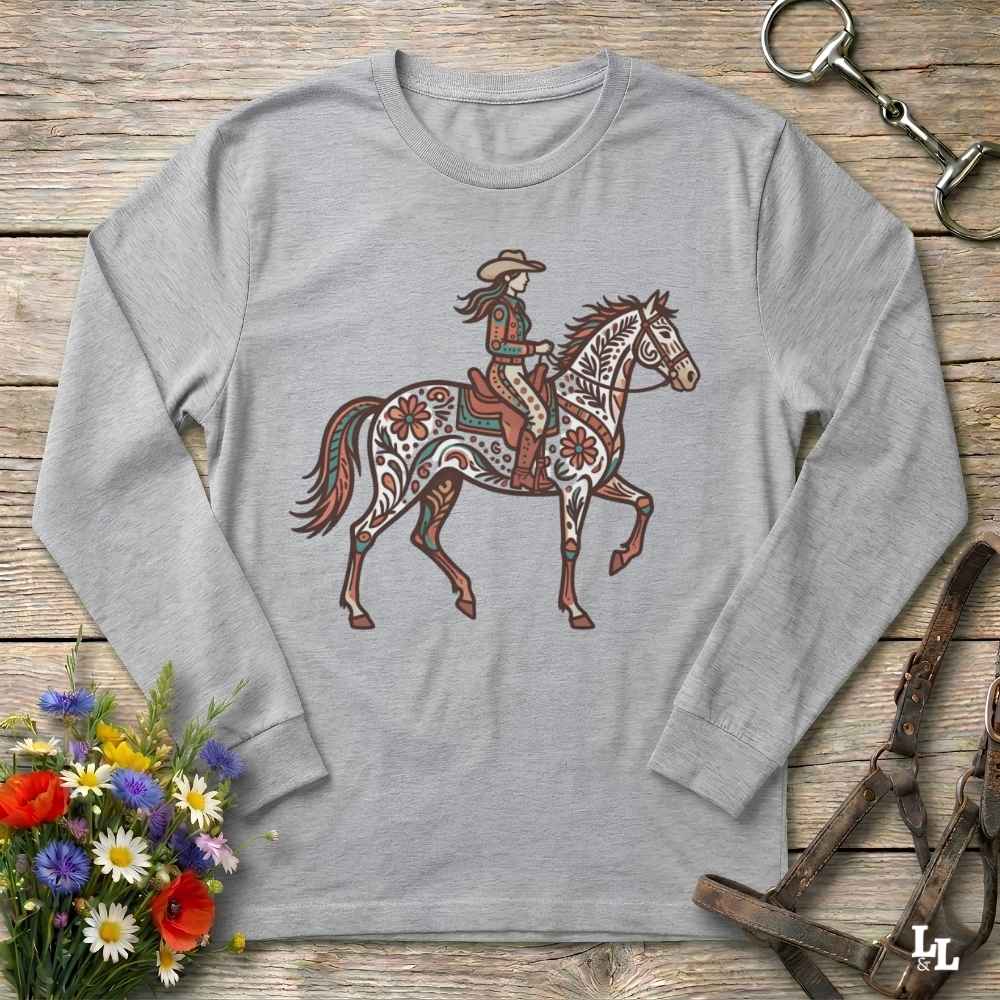 A grey long-sleeve t-shirt with a western-themed graphic of a rider on a decorated horse, displayed on a rustic wooden background with equestrian accessories and flowers.