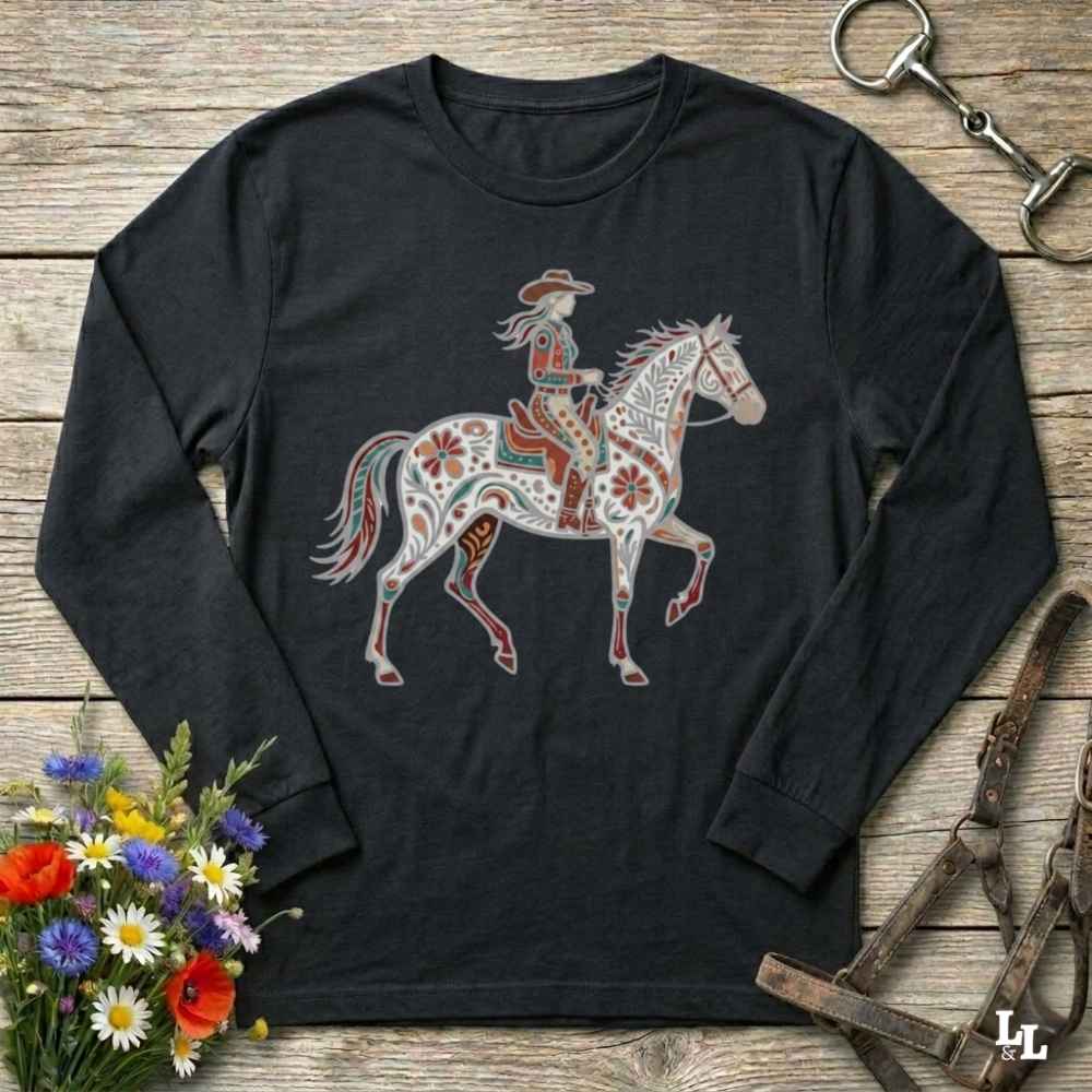 A black long-sleeve t-shirt with a western-themed graphic of a rider on a decorated horse, displayed on a rustic wooden background with equestrian accessories and flowers.