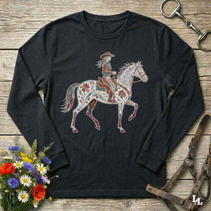 A black long-sleeve t-shirt with a western-themed graphic of a rider on a decorated horse, displayed on a rustic wooden background with equestrian accessories and flowers.