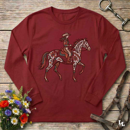 A long-sleeve crewneck t-shirt in maroon red, featuring a graphic of a cowboy riding a decorated horse. The shirt is displayed flat on a rustic wooden background with equestrian-themed props like a leather bridle and a bouquet of wildflowers.