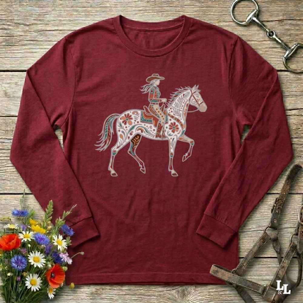 Maroon long-sleeve shirt with a colorful horse and rider design on a wooden surface with flowers and equestrian equipment.