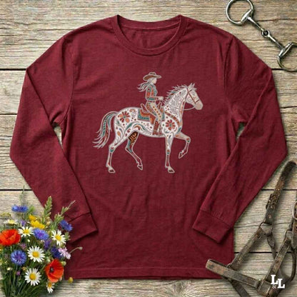 Maroon long-sleeve shirt with a colorful horse and rider design on a wooden surface with flowers and equestrian equipment.