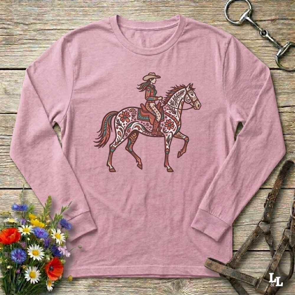 Pink long-sleeve shirt with a graphic of a woman riding a horse on a wooden surface with floral and equestrian-themed accessories.
