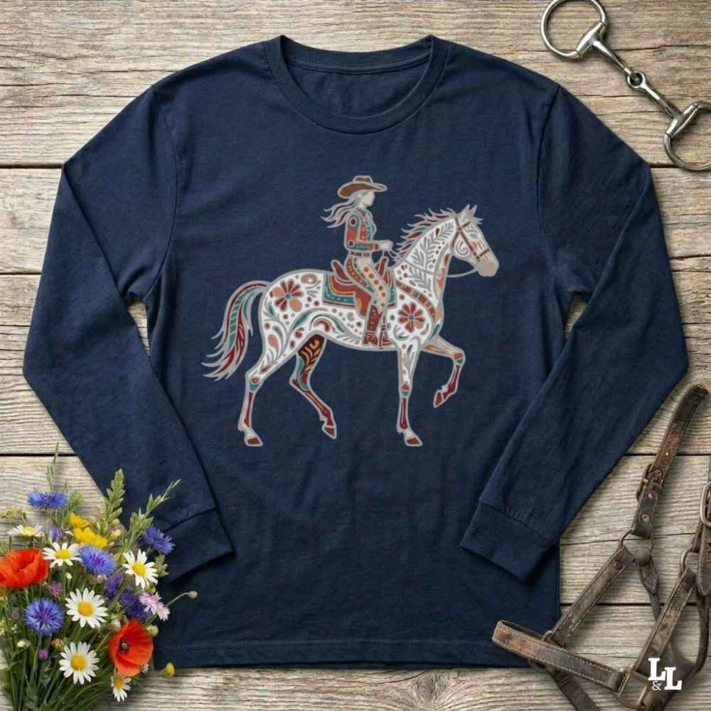 A navy blue long-sleeve t-shirt with a western-themed graphic of a cowboy riding a decorated horse, displayed on a rustic wooden background with equestrian accessories and flowers.