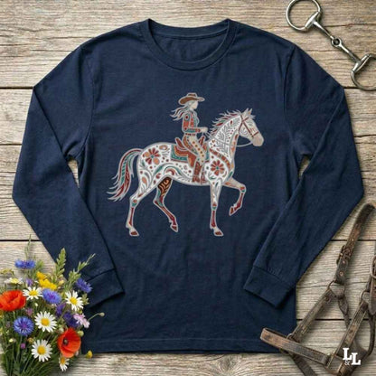 A navy blue long-sleeve t-shirt with a western-themed graphic of a cowboy riding a decorated horse, displayed on a rustic wooden background with equestrian accessories and flowers.
