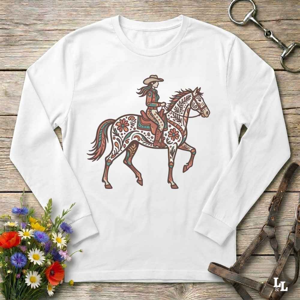 A white long-sleeve t-shirt with a western-themed graphic of a rider on a decorated horse, displayed on a rustic wooden background with equestrian accessories and flowers.