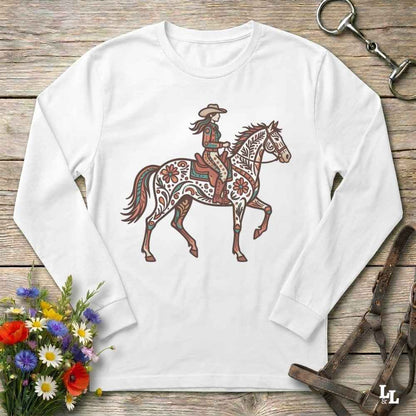 A white long-sleeve t-shirt with a western-themed graphic of a rider on a decorated horse, displayed on a rustic wooden background with equestrian accessories and flowers.