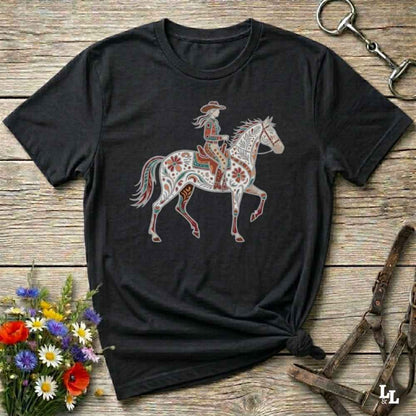 Black t-shirt with a colorful horse and rider graphic on a wooden surface with floral and equestrian-themed accessories.