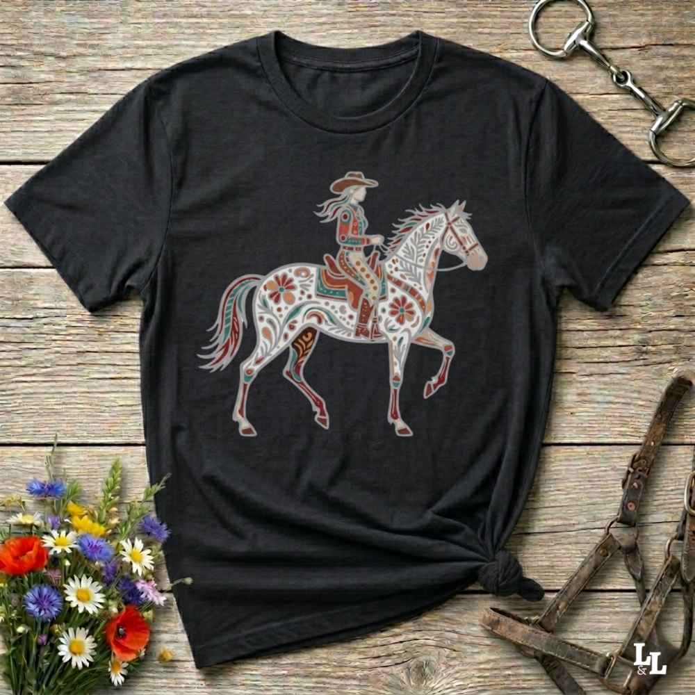 A black short-sleeve t-shirt with a colorful graphic of a cowboy riding a decorated horse, styled on a rustic wooden background with flowers and horse tack.