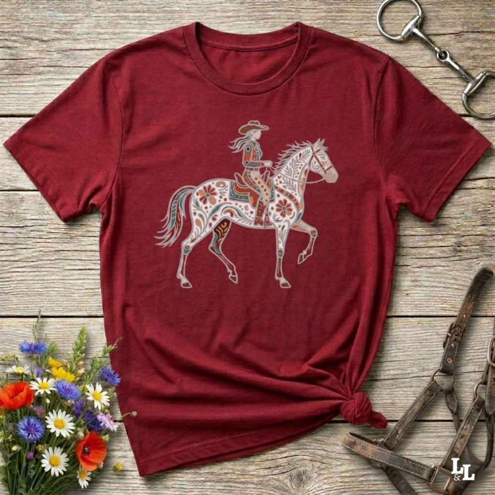 Red t-shirt with a colorful graphic of a woman on a horse, placed on a wooden surface with floral and equestrian-themed accessories.