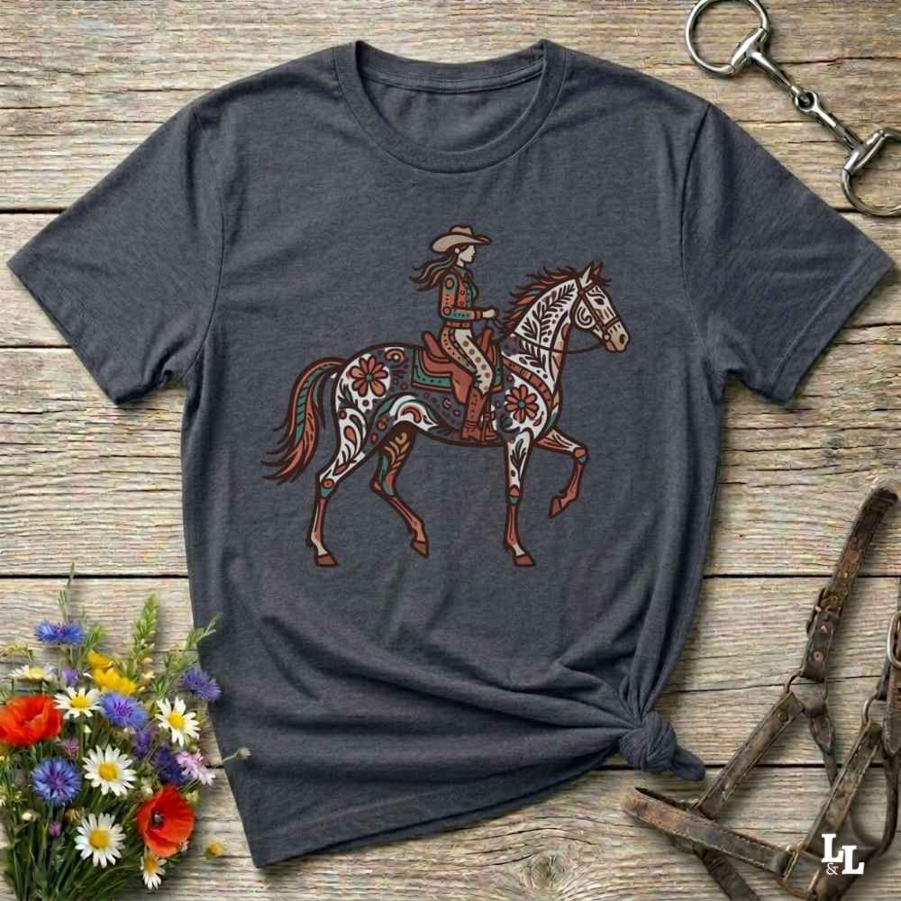 A dark heather gray t-shirt with a western-themed graphic of a decorated cowboy riding a horse, styled in a flat-lay on a rustic wooden background with equestrian accessories and flowers.