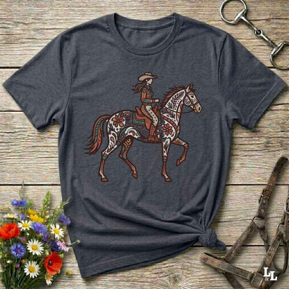 A dark heather gray t-shirt with a western-themed graphic of a decorated cowboy riding a horse, styled in a flat-lay on a rustic wooden background with equestrian accessories and flowers.