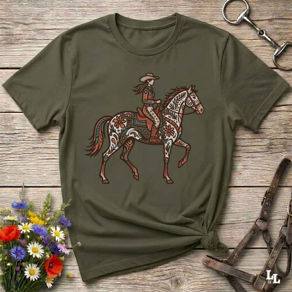 An olive green short-sleeve t-shirt with a graphic of a cowboy riding a decorated horse, laid flat on a rustic wooden surface with floral and equestrian props.